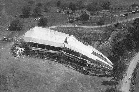 A wrecked part of an airship lays in a field with steel skeleton exposed, resting near a line of trees